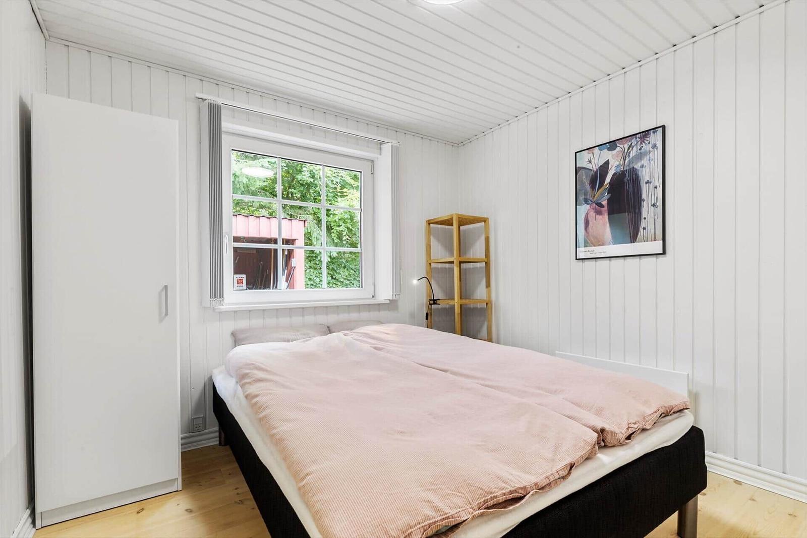 Schlafzimmer mit Bett, Kleiderschrank und Fenster. Wandbild und Holzregal sind sichtbar.