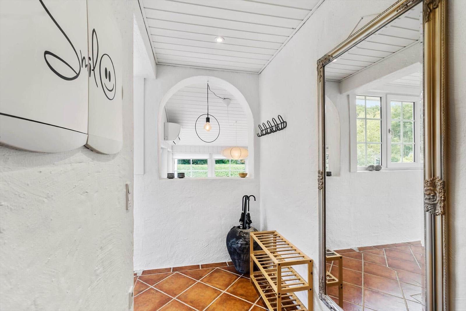Eingangsbereich mit Spiegel, Holzständer und Terrakottafußboden. Fenster und Decke sind weiß.