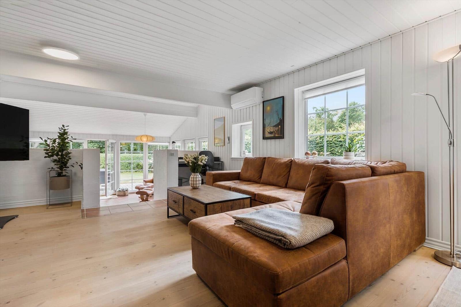 Wohnzimmer mit braunem Sofa, Holzboden und weißen Wänden. Fenster mit Blick auf Garten.