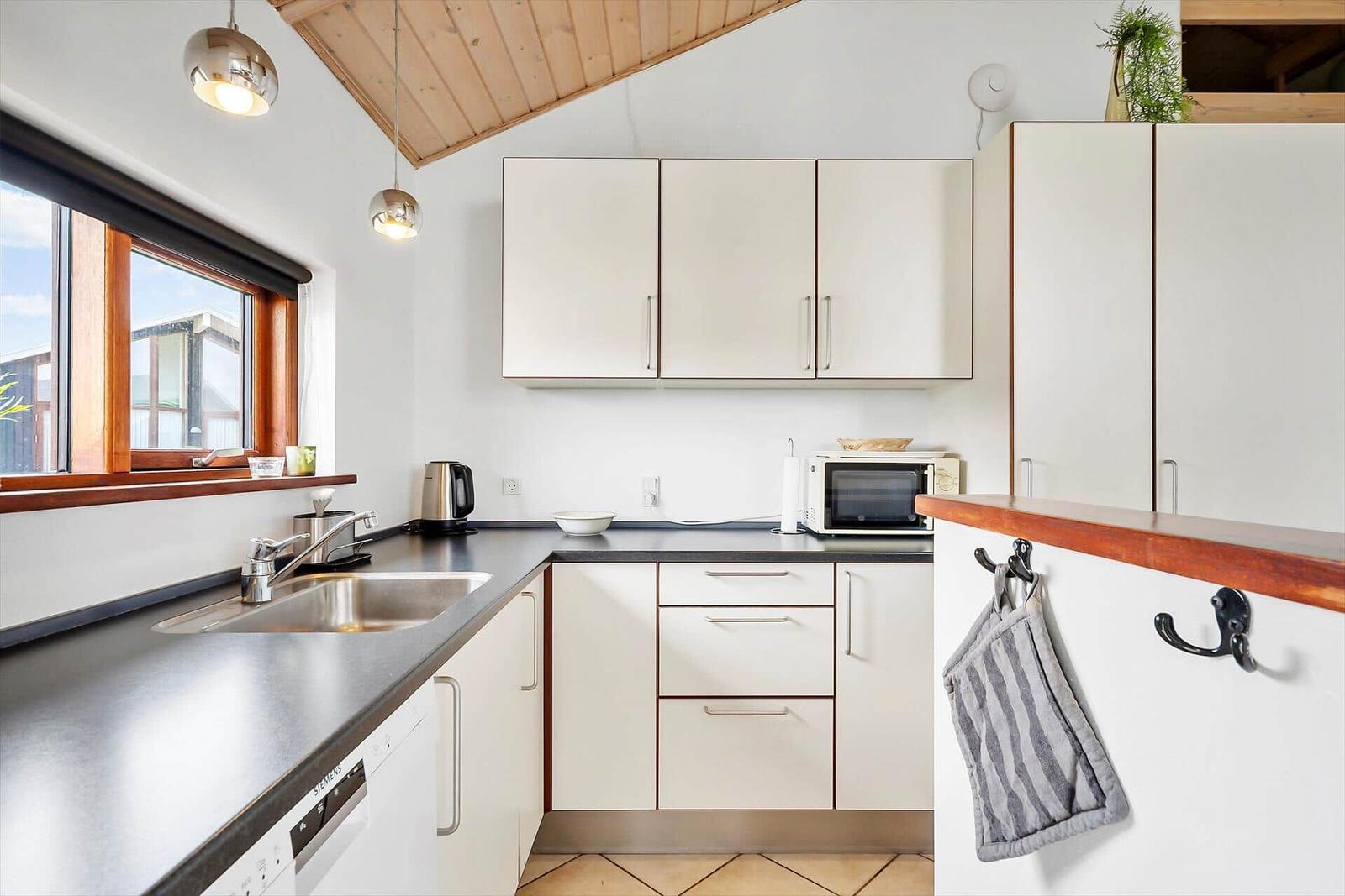 Küche mit weißen Schränken, Edelstahlspüle und Mikrowelle. Holzfenster und Decke.
