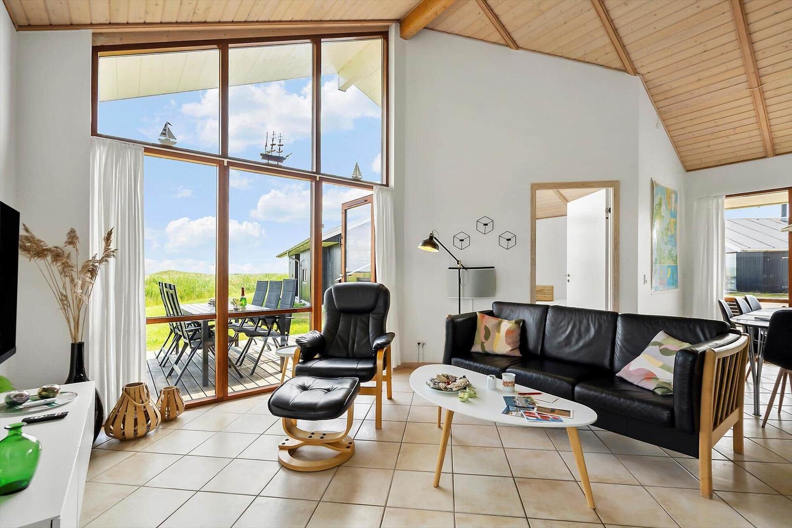 Wohnraum mit schwarzer Ledercouch und Sessel, großes Fenster mit Blick auf Terrasse und Landschaft.