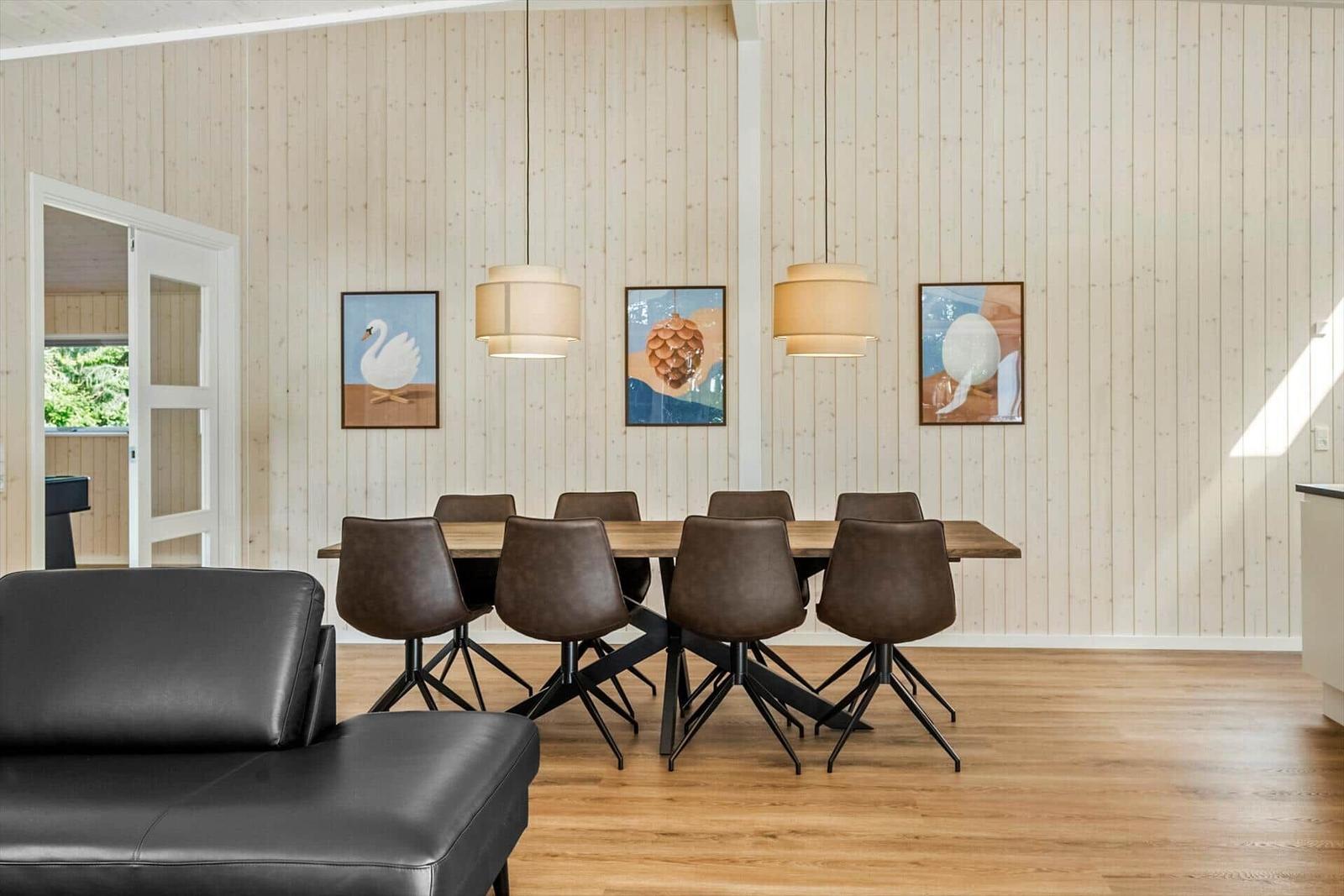 Dining room with wooden wall, table, six chairs, three wall pictures, and two pendant lights.