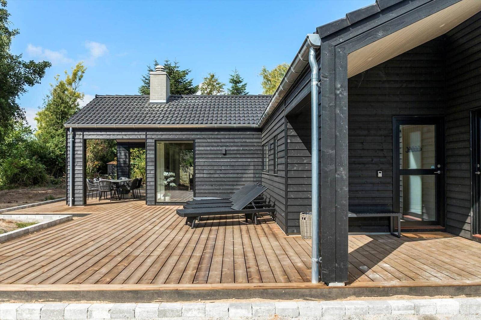 Modern villa with wooden terrace, lounge chairs, and garden furniture. Black facade and large outdoor area.