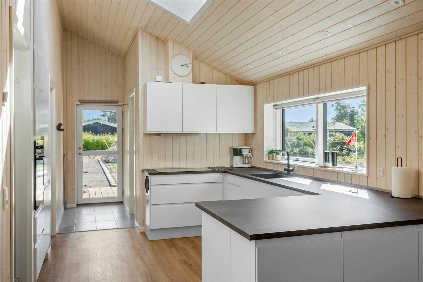 Modern kitchen with white cabinets and dark worktop. Wooden walls and large windows.