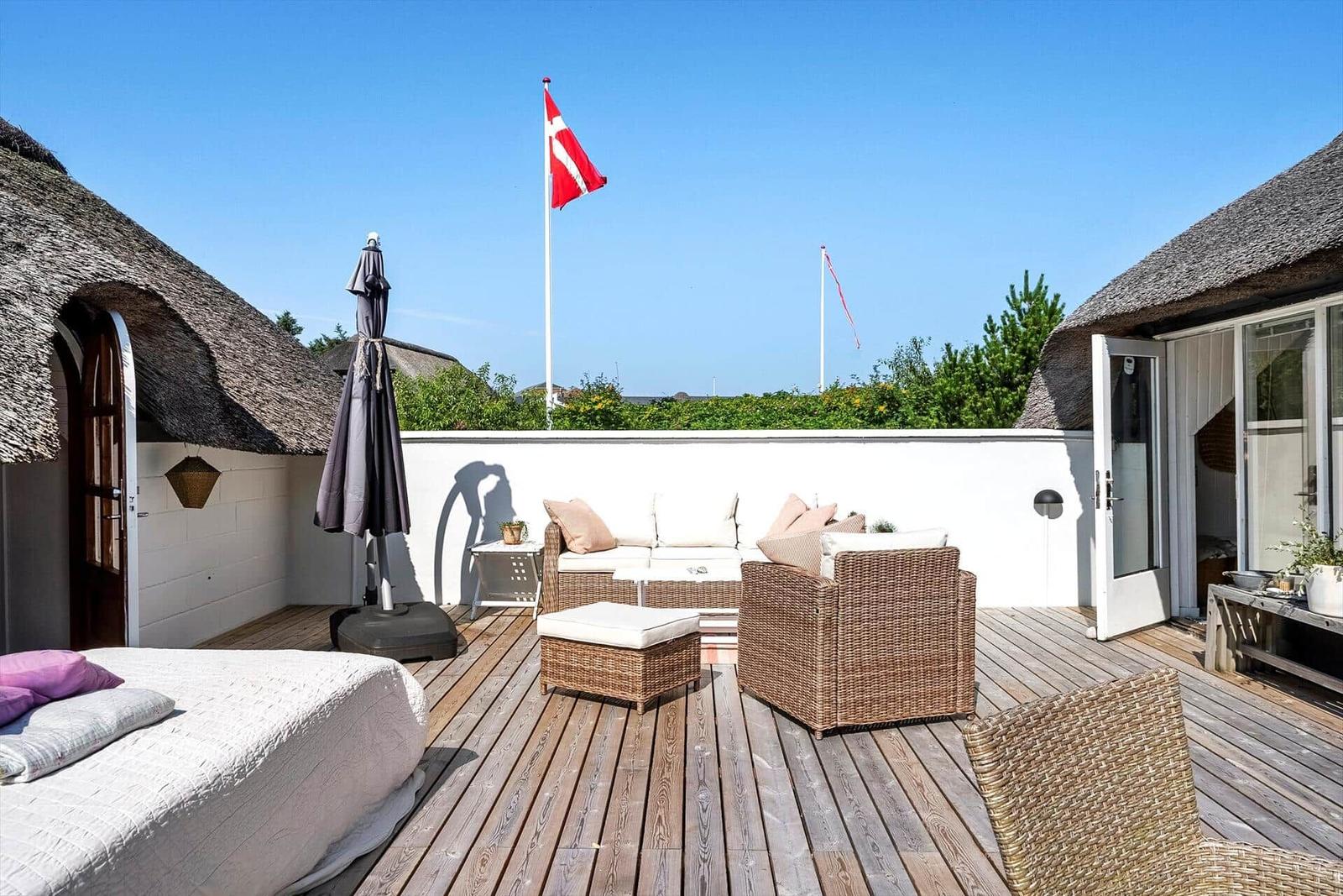 Terrasse mit Liege, Sitzgruppe und dänischer Flagge. Holzdecke und Strohdach.