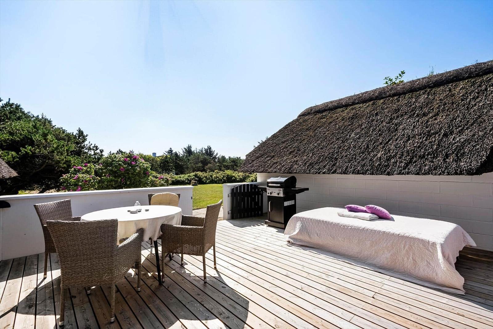 Terrasse mit Tisch, Stühlen, Grill und Liege unter strohgedecktem Dach.