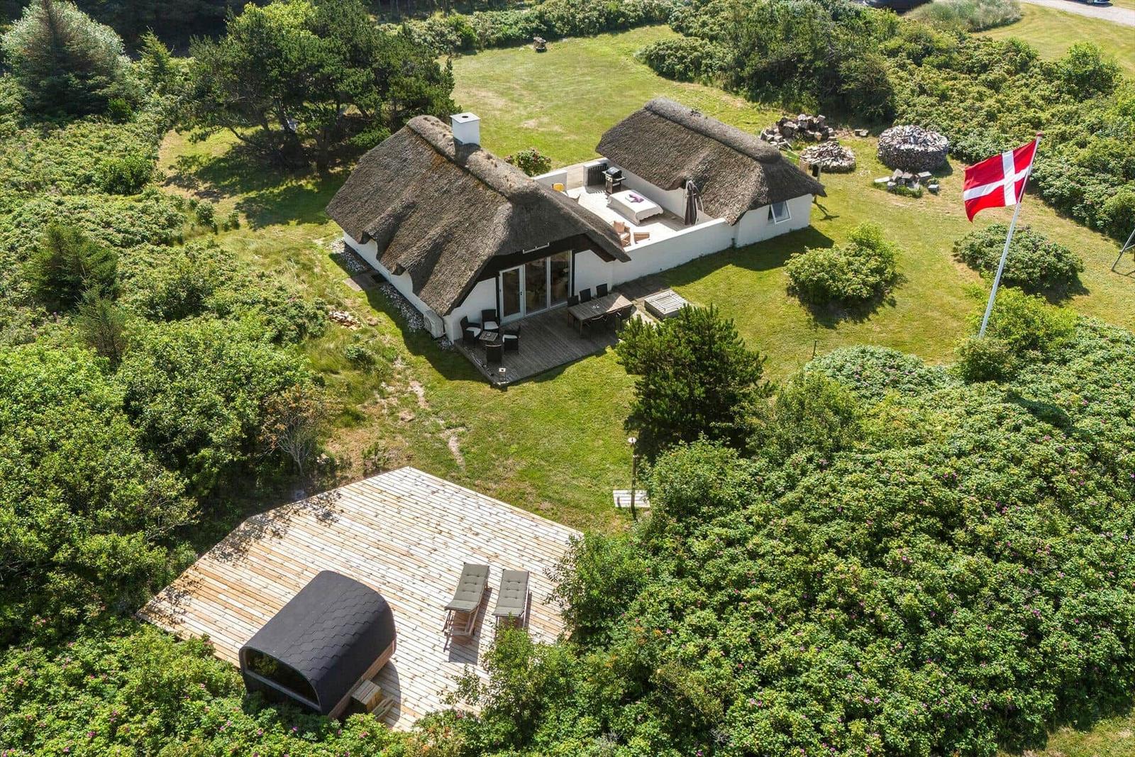 Dänische Ferienhaus mit Strohdach, Terrasse und Garten. Daneben steht eine moderne Sauna auf Holzterrasse.