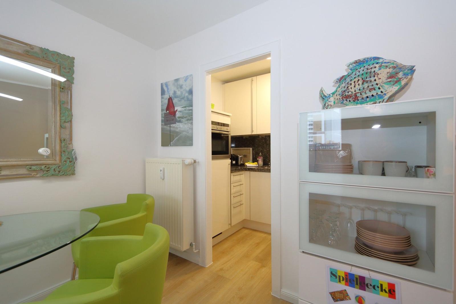 Essbereich mit grünen Stühlen und Glasplatte. Küche im Hintergrund. Glasvitrine mit Geschirr.