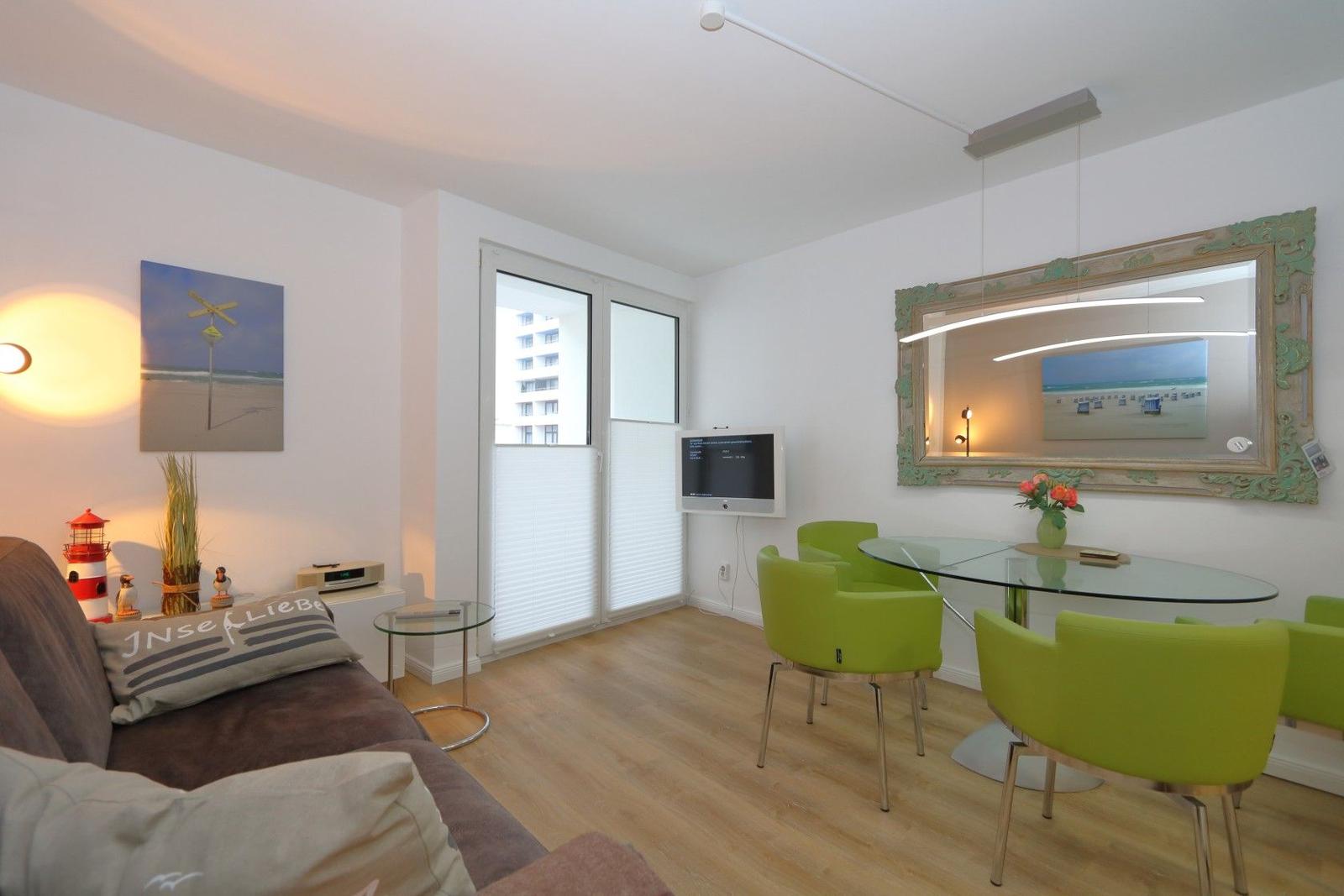 Wohnzimmer mit Esstisch, grünen Stühlen und Fernseher an der Wand.