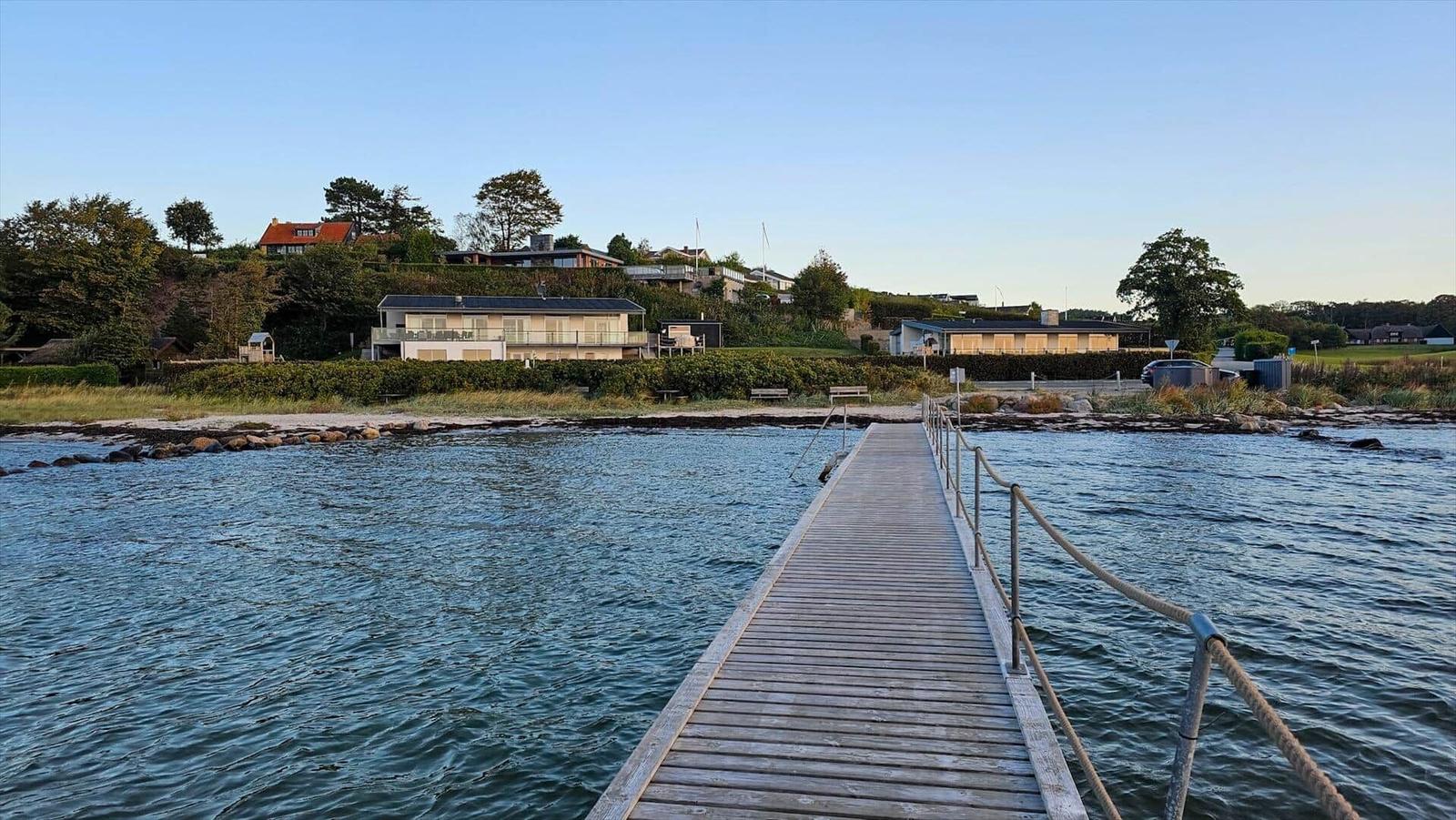 Holzsteg führt über Wasser zu Strandhaus mit Blick auf grüne Hügel.
