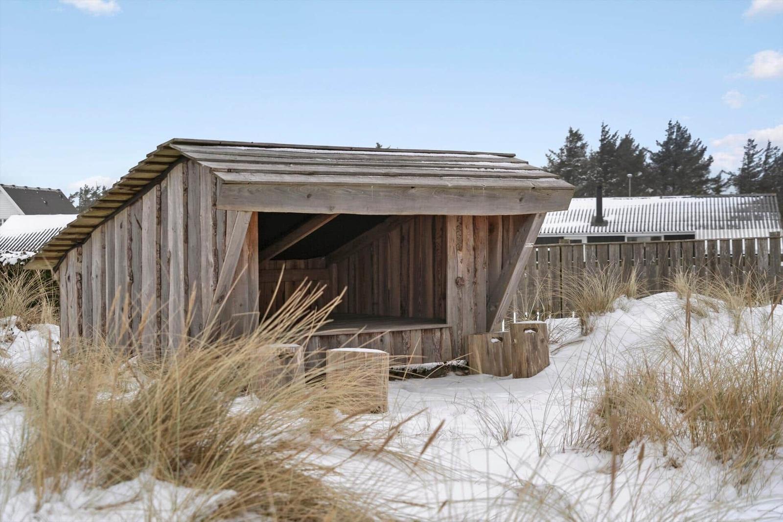 Holzschuppen mit Sitzbank im Schnee, umgeben von Gräsern und Bäumen.