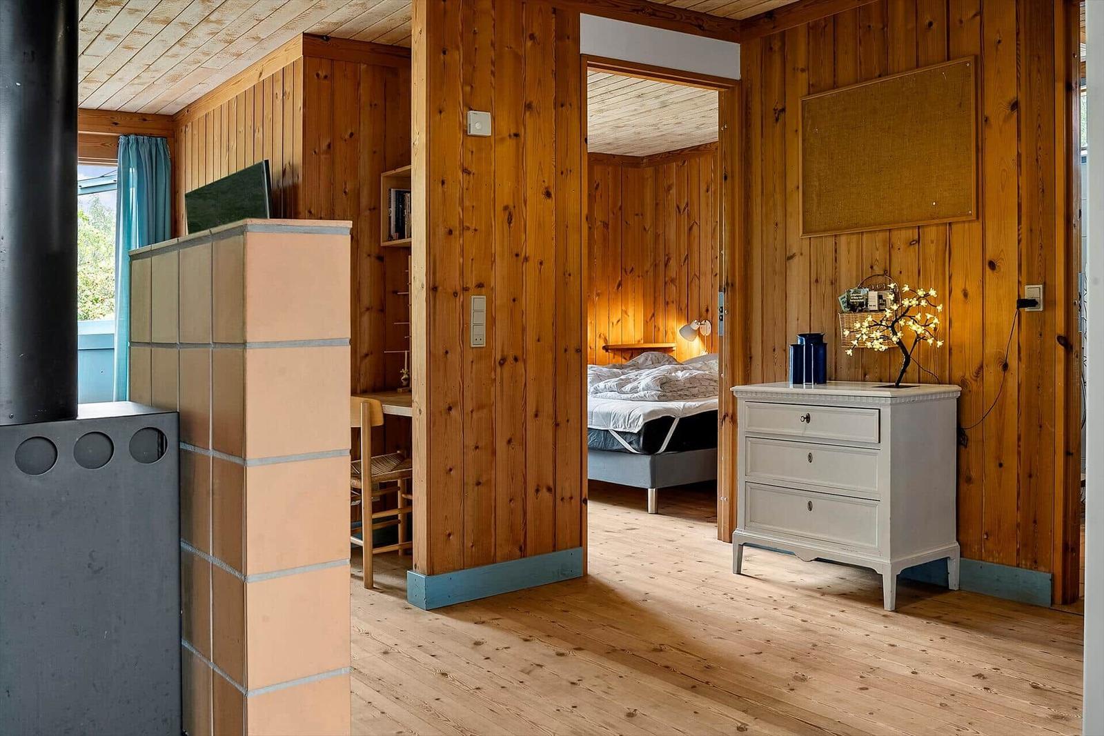 Living area with wooden walls and bedroom in background. White dresser and tiled wall visible.