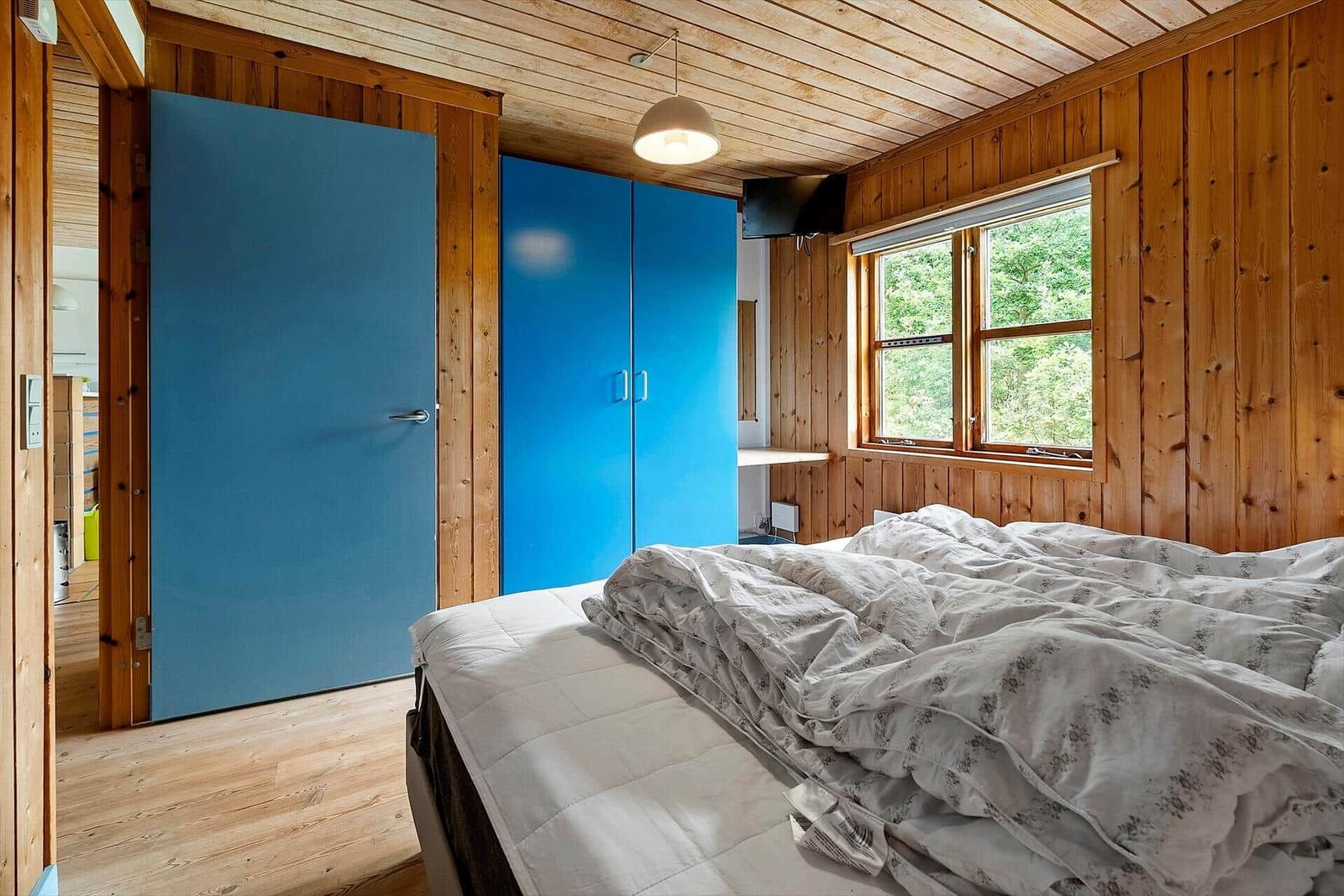 Bedroom with wooden walls, blue door and wardrobe, bed with patterned bedding and window with view of forest.