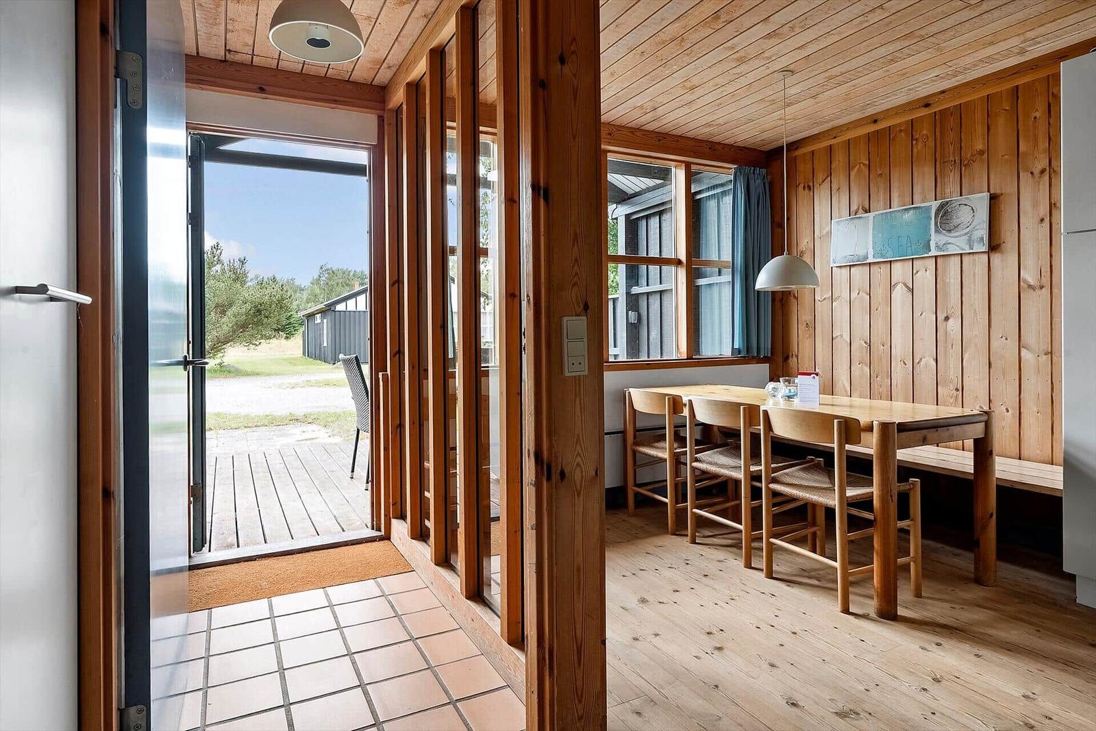 Dining area with wooden table, chairs, and windows. Passage to terrace with wooden planks.