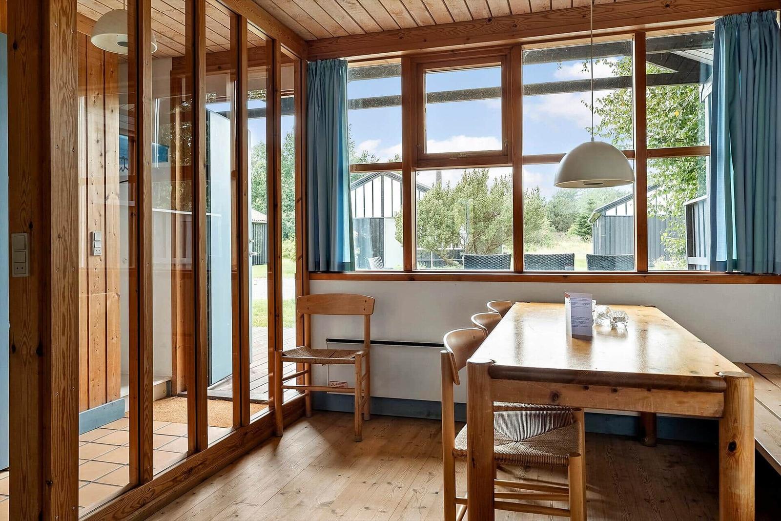 Dining room with wooden table, chairs, and large windows. View of garden and adjacent buildings.