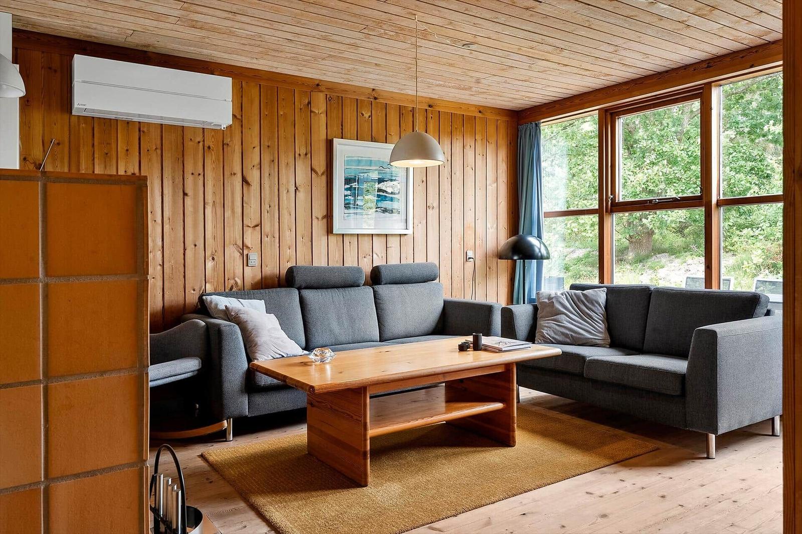 Living room with wooden wall, two gray sofas, and wooden table. Large windows with view of forest.