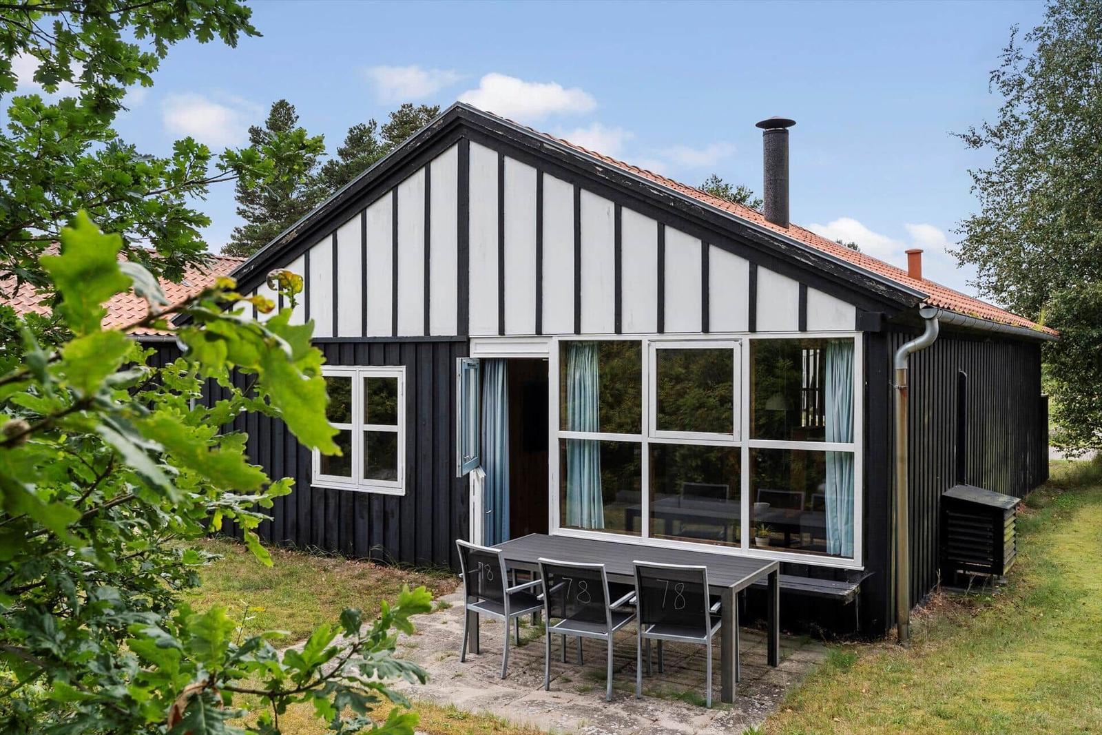 Black wooden house with white timber frame, terrace with table and chairs, surrounded by trees.