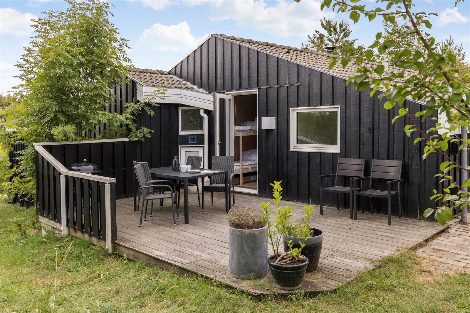 Schwarze Holzhütte mit Terrasse, Tisch, Stühlen und Grill. Im Hintergrund sind Bäume und ein Garten.
