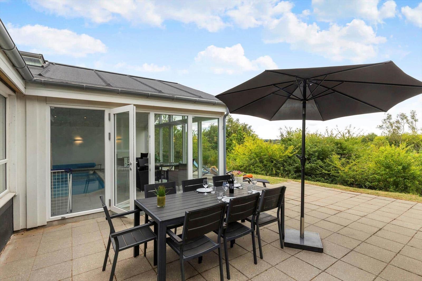 Terrasse mit Tisch, Stühlen und Sonnenschirm. Durchgehende Glasfront zum Poolbereich.