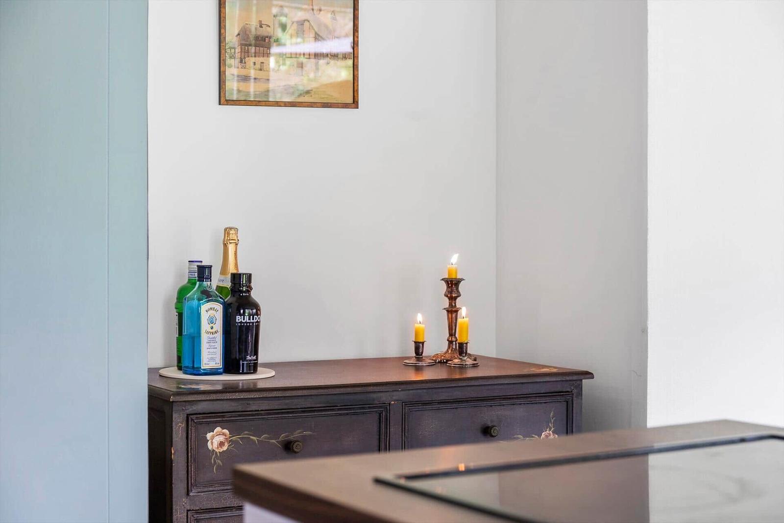 Wooden cabinet with bottles and lit candles. Wall picture above the cabinet.