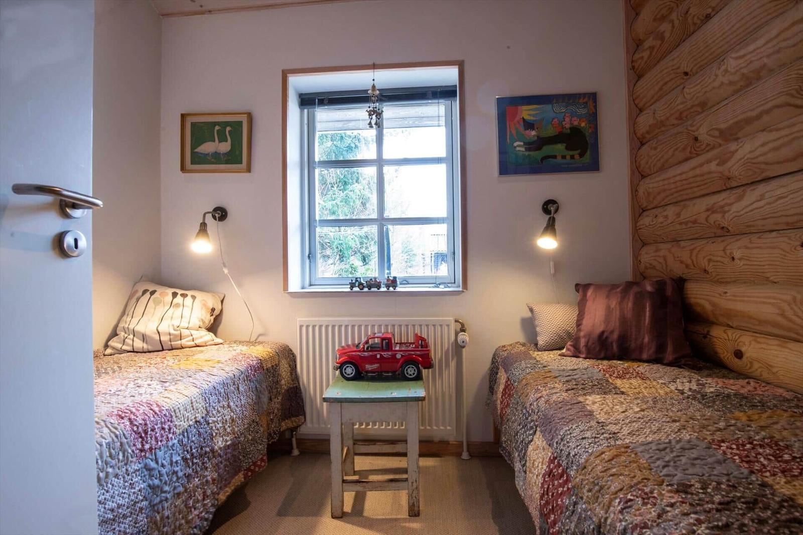 Zwei Betten mit Quiltdecken stehen im Raum. Fenster, Wandlampen und Holzwand sind sichtbar.