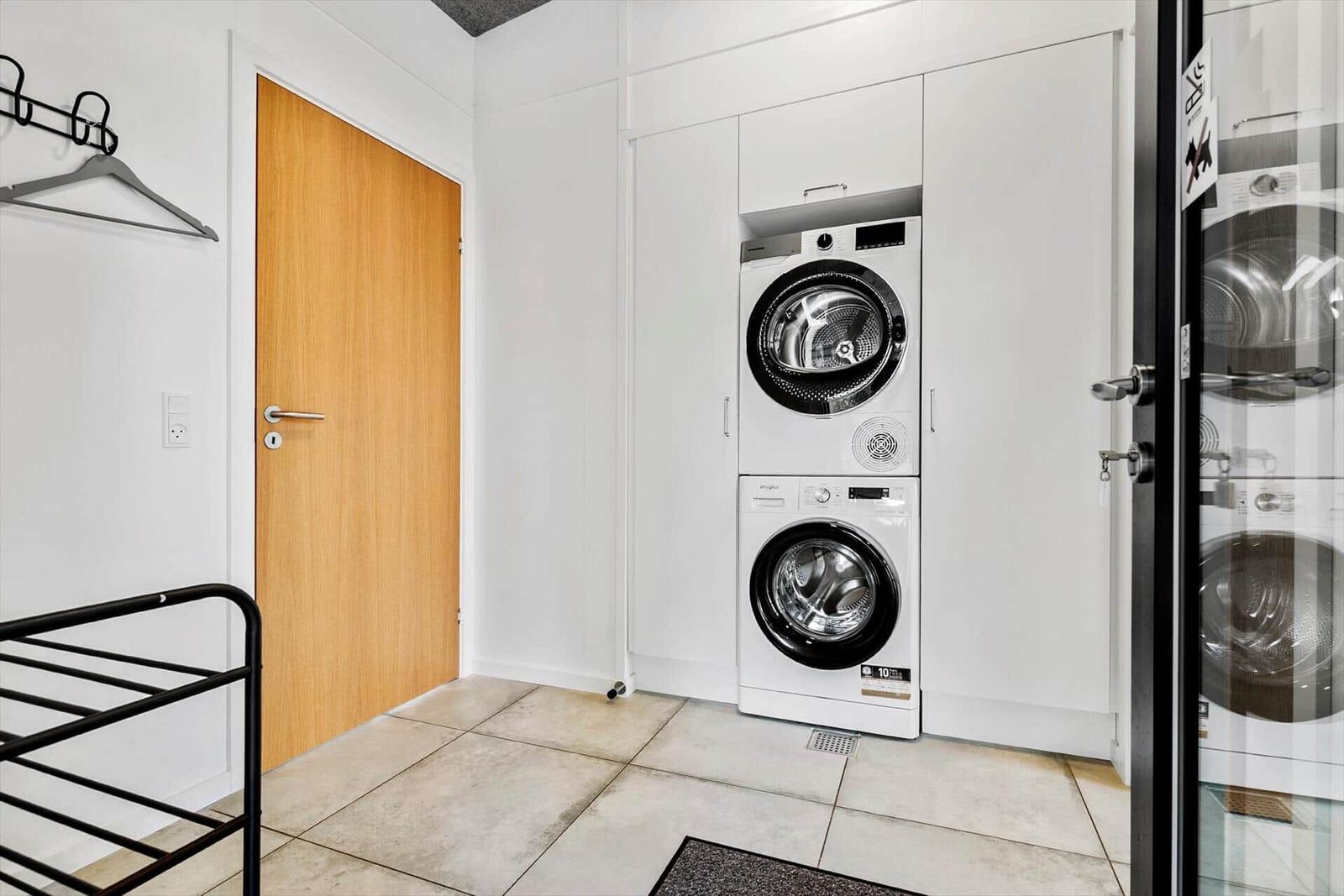 Waschmaschine und Trockner in weißer Einbauküche mit Holztür und schwarzer Garderobe.