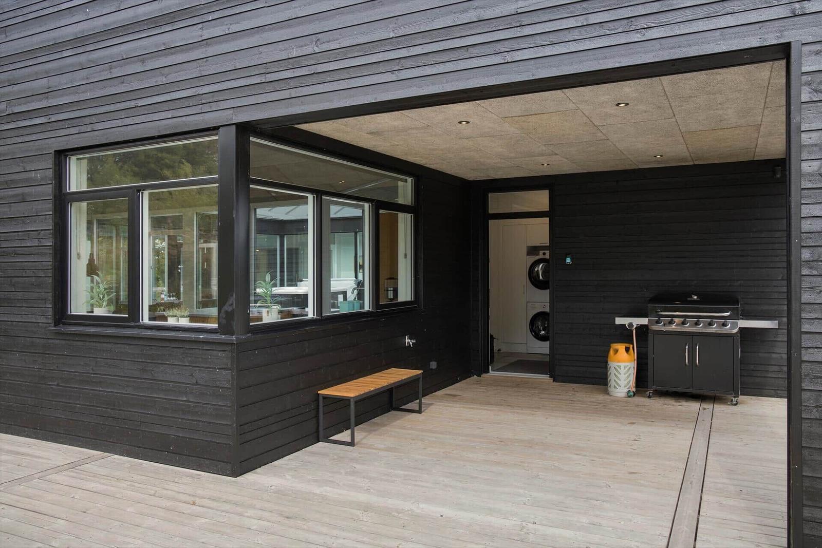 Terrasse mit Grill, Waschmaschine und Holzbänkchen an schwarzem Holzhaus.