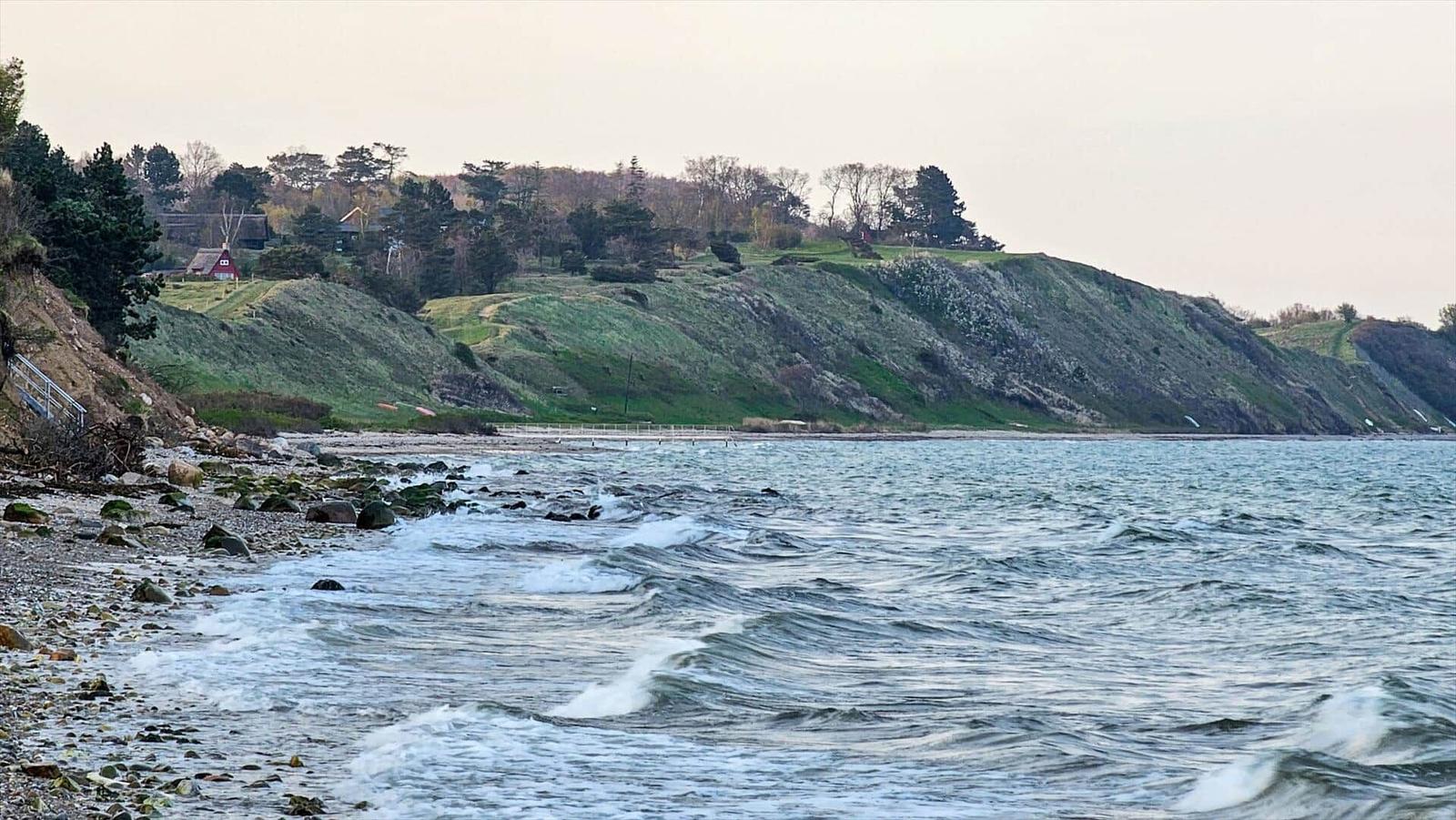 Beach with rocks and waves, behind green hills with trees and a red house.