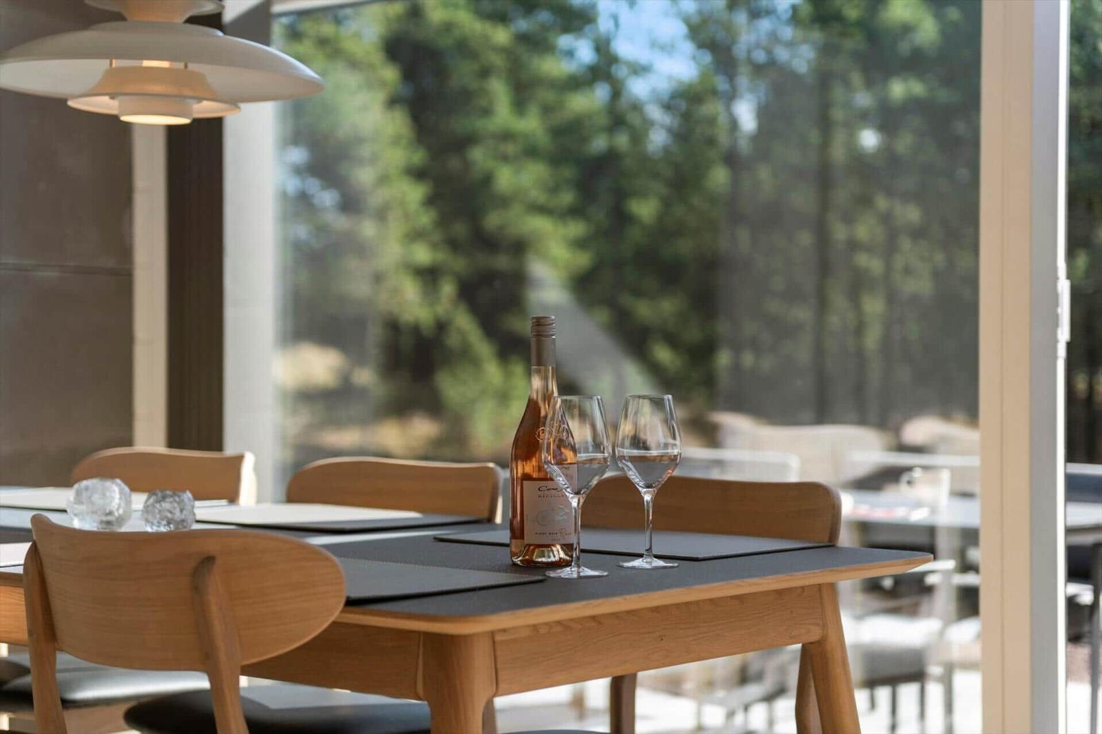 Dining table with wine bottle and glasses in front of large window with garden view.
