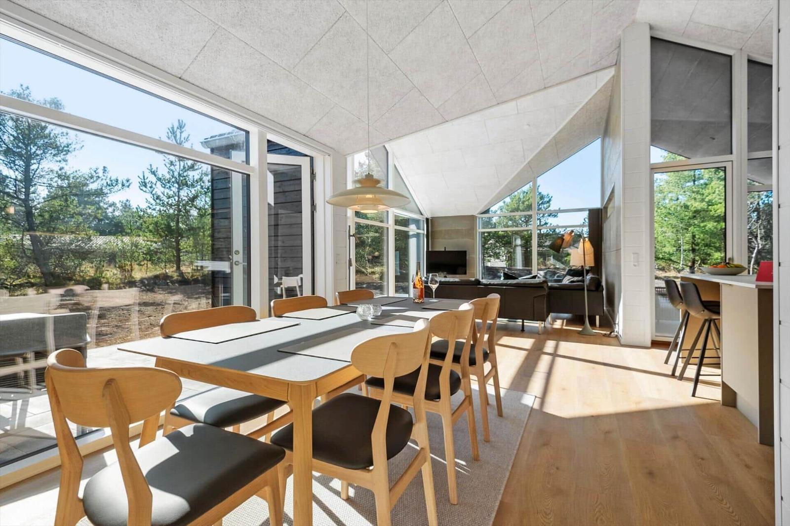 Dining area with wooden table and chairs. Large windows show forest. Living area with sofa and TV.