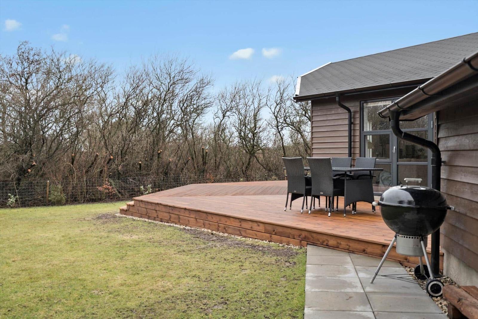 Wooden terrace with seating and grill beside house. Lawn and trees in background.