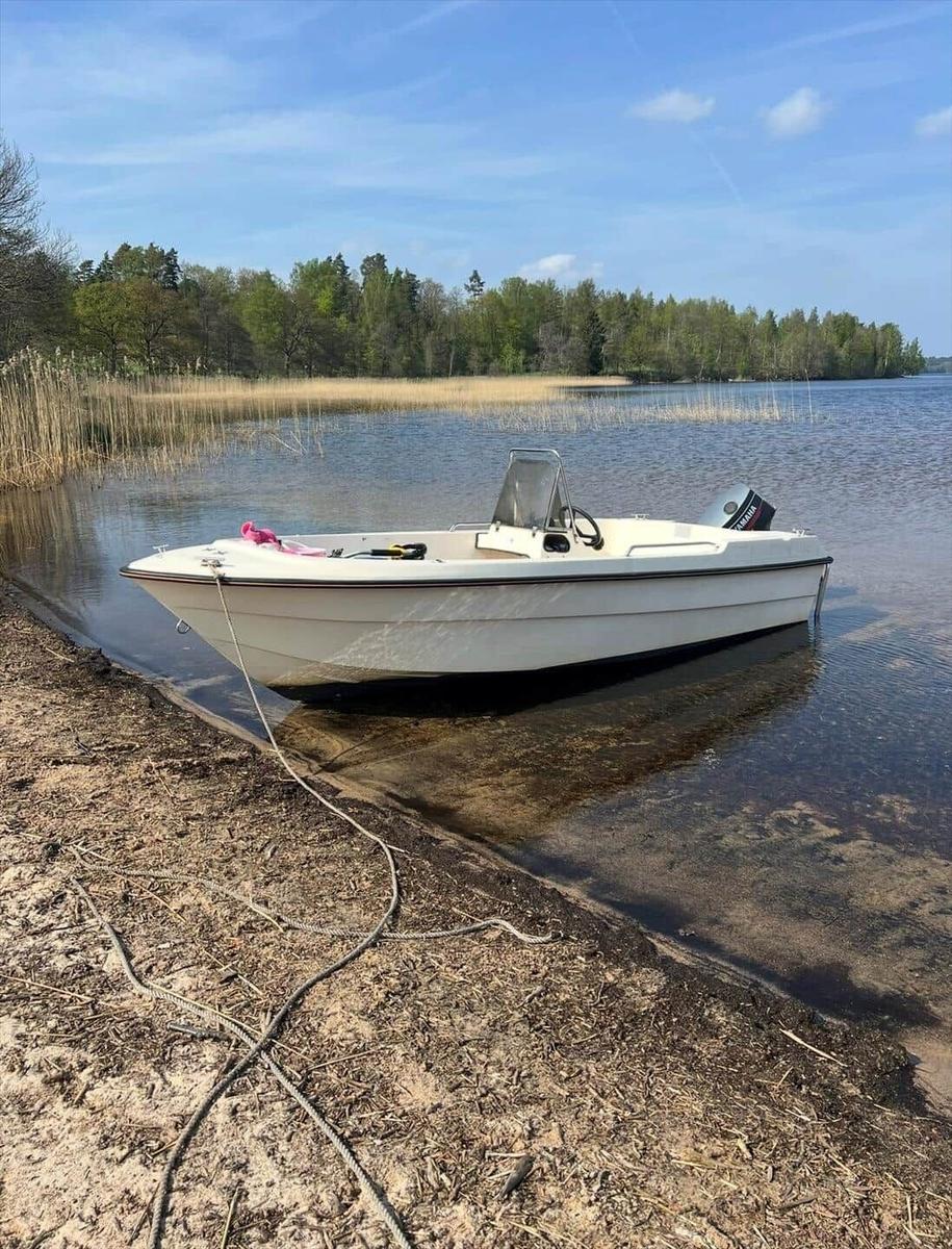 Ein weißes Motorboot liegt am Ufer eines Sees mit Gras und Bäumen im Hintergrund.
