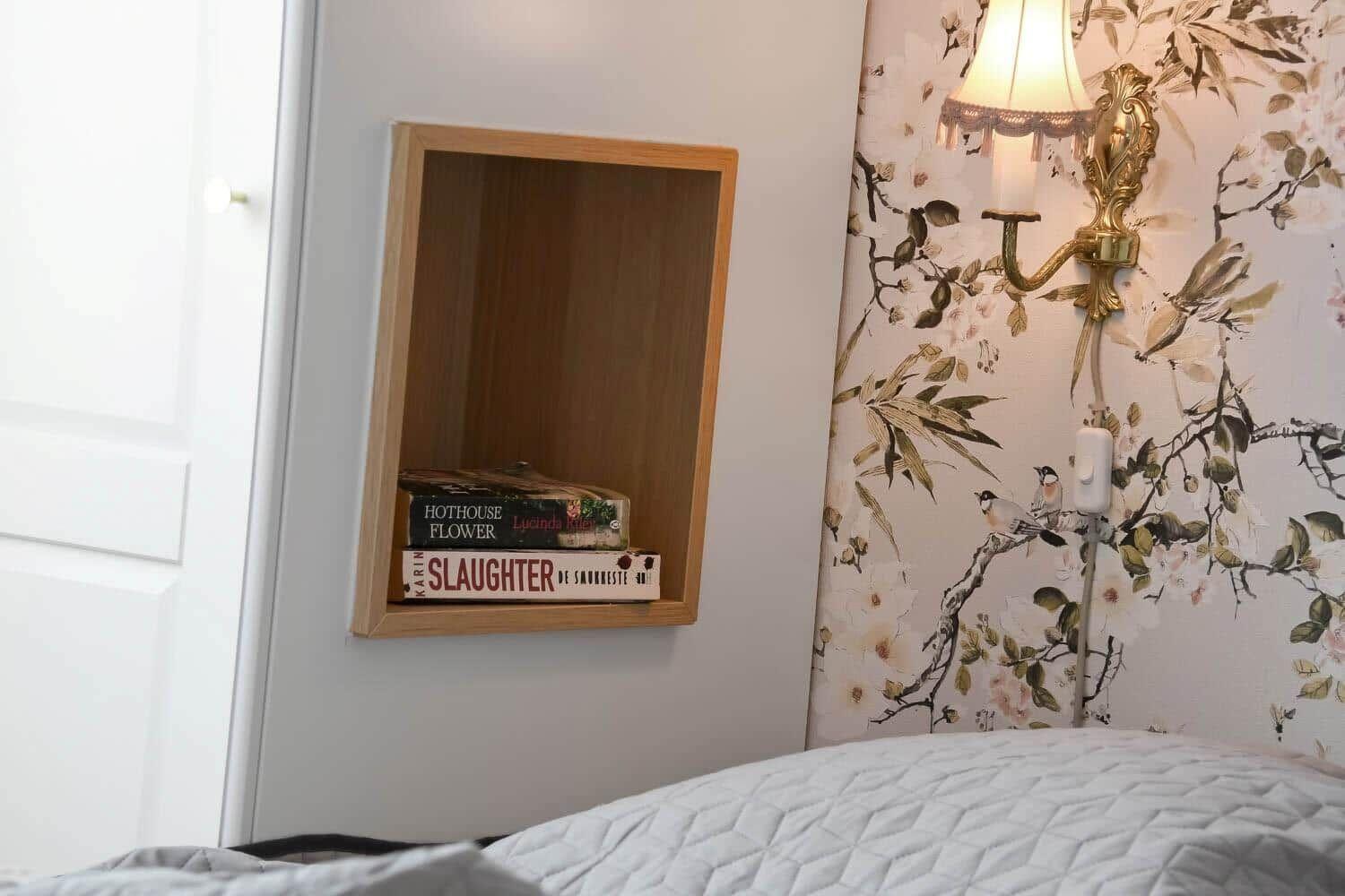 Bettkante mit Holzregal, Blumenwand und Wandlampe.