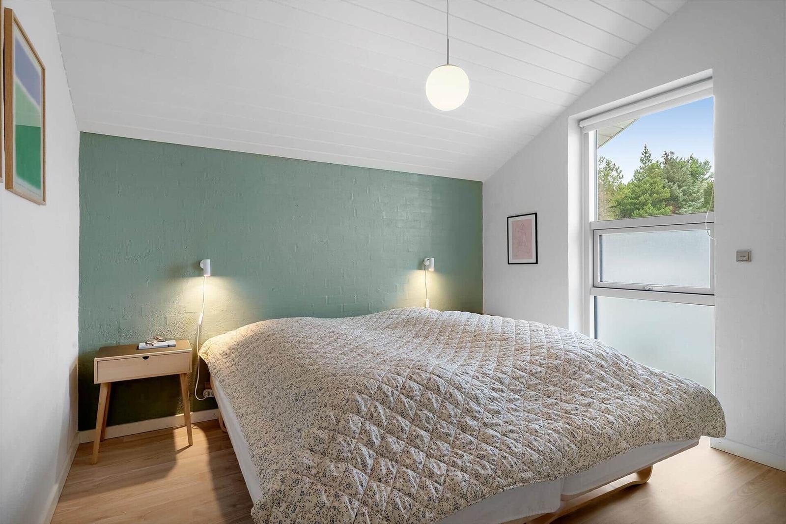 Schlafzimmer mit Bett, Nachttisch und Fenster. Wand in Grün, Holzboden und Deckenleuchte.