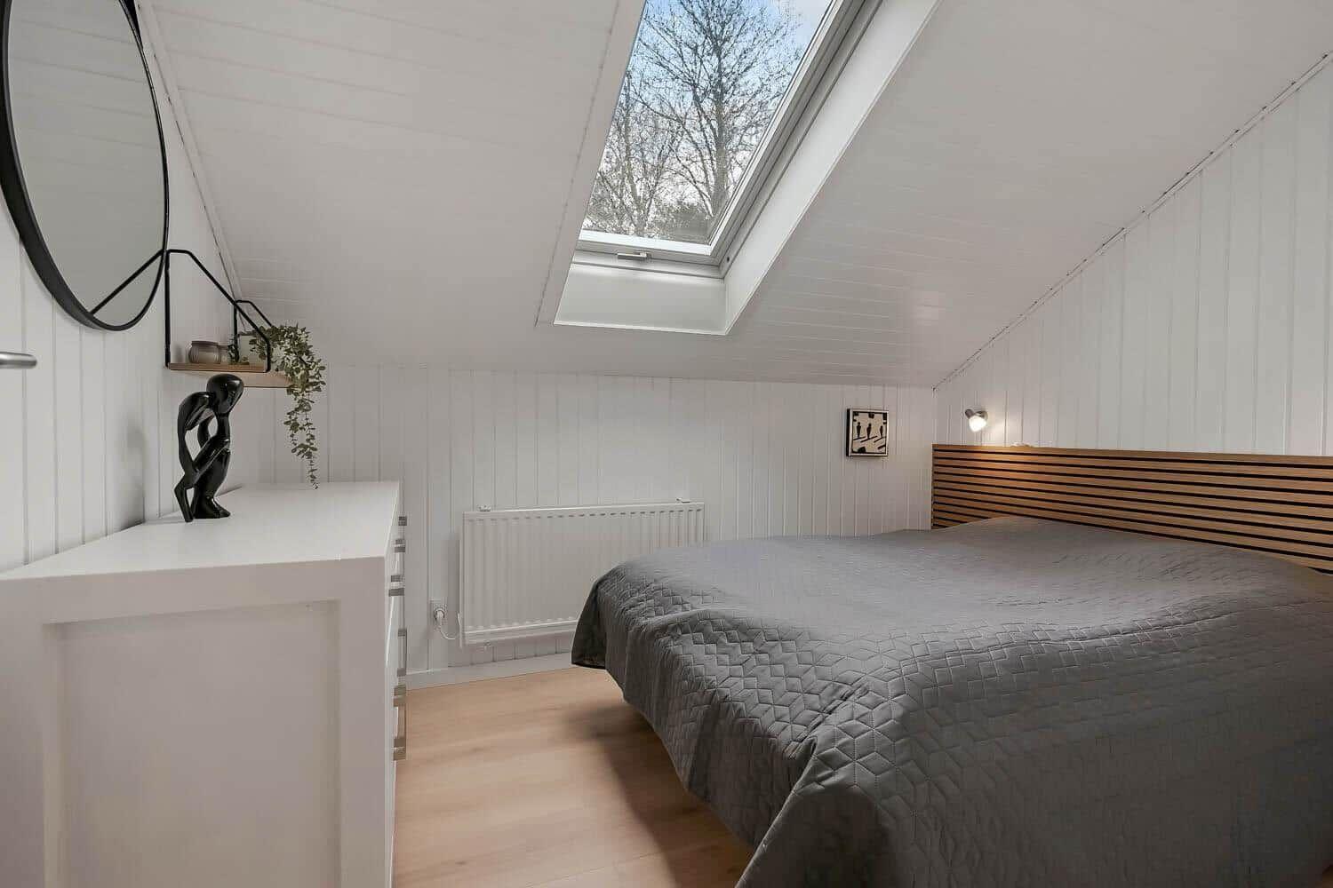 Schlafzimmer mit Dachschrägen, Dachfenster und Holzboden. Bett mit grauer Bettwäsche und weißer Kommode.