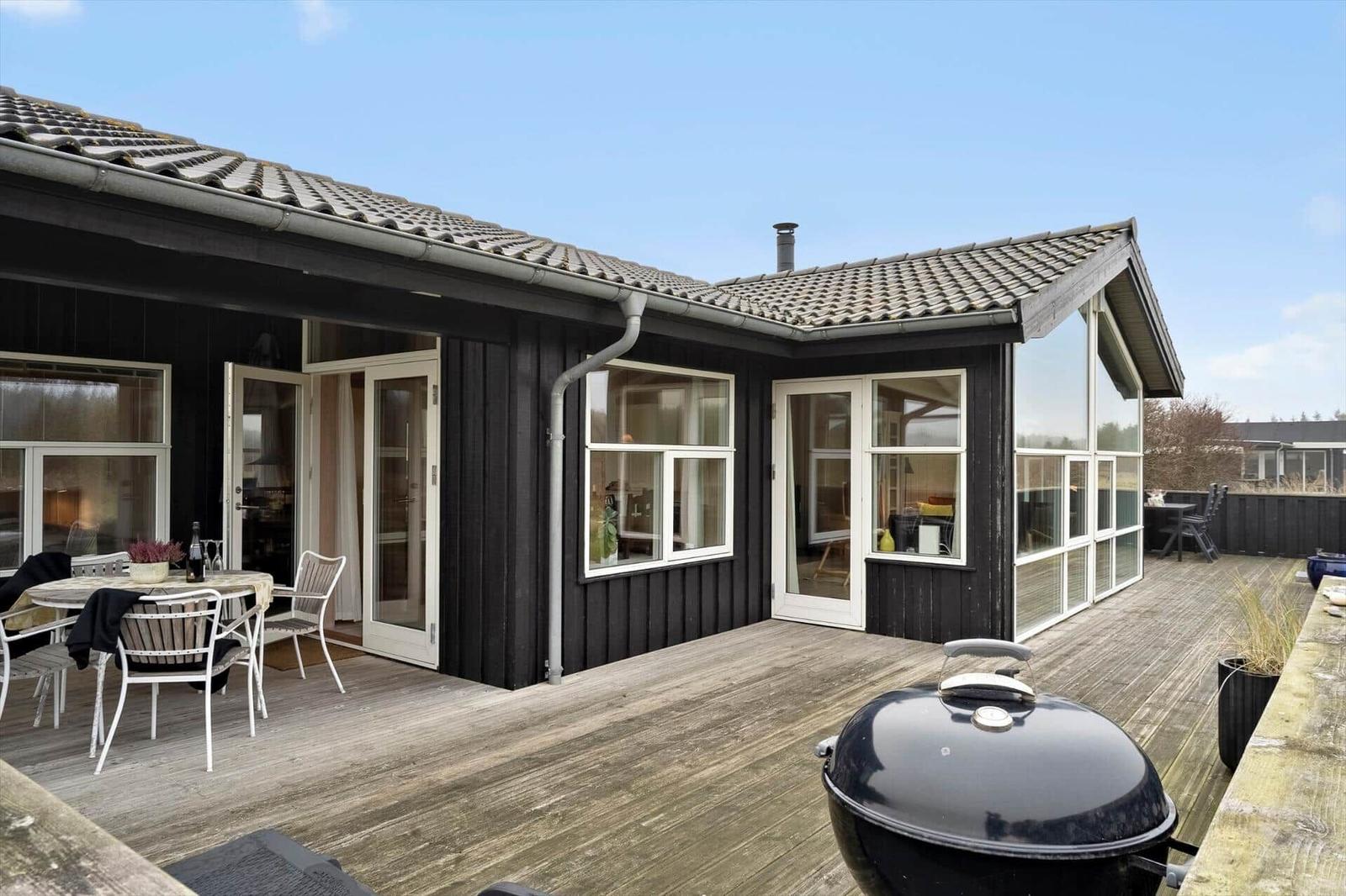Haus mit Holzterrasse, Grill und Sitzgelegenheiten. Fenster und Türen zeigen Innenräume.