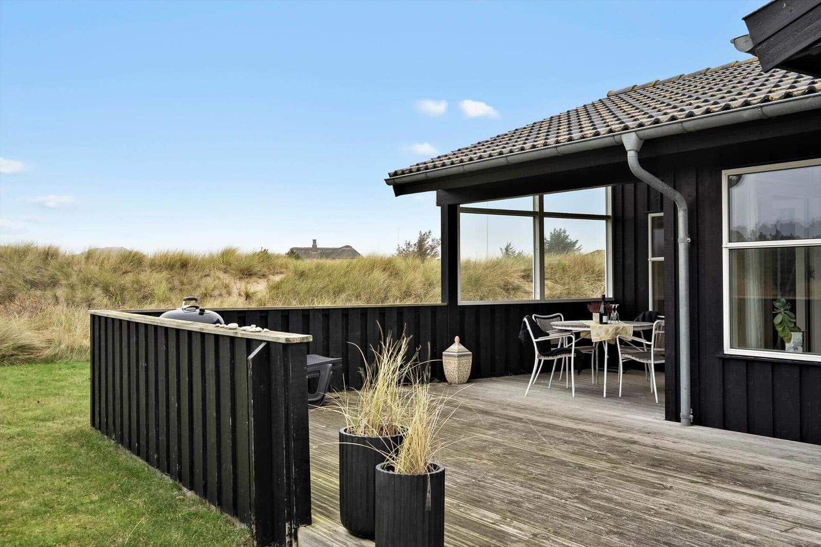 Terrasse mit Tisch, Stühlen und Grill. Schwarze Holzwand, Pflanzen und Dachüberstand.