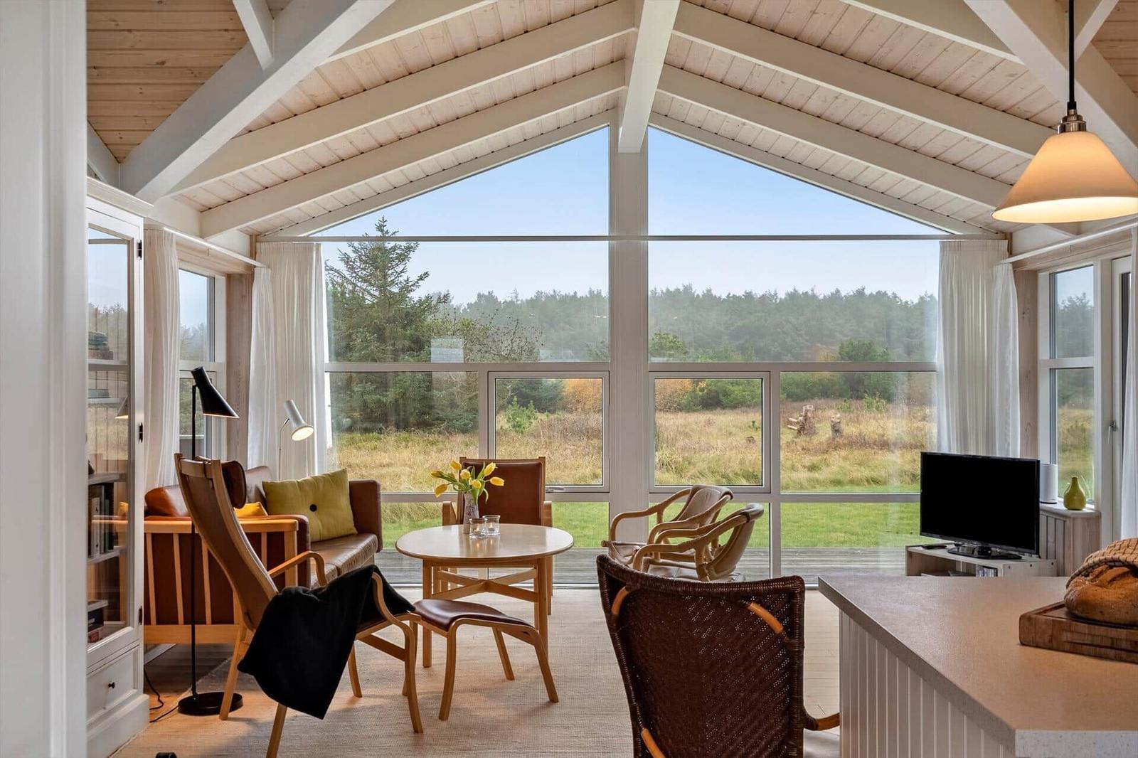 Großes Wohnzimmer mit Blick auf Wald. Holzdecke, große Fenster, Möbel.