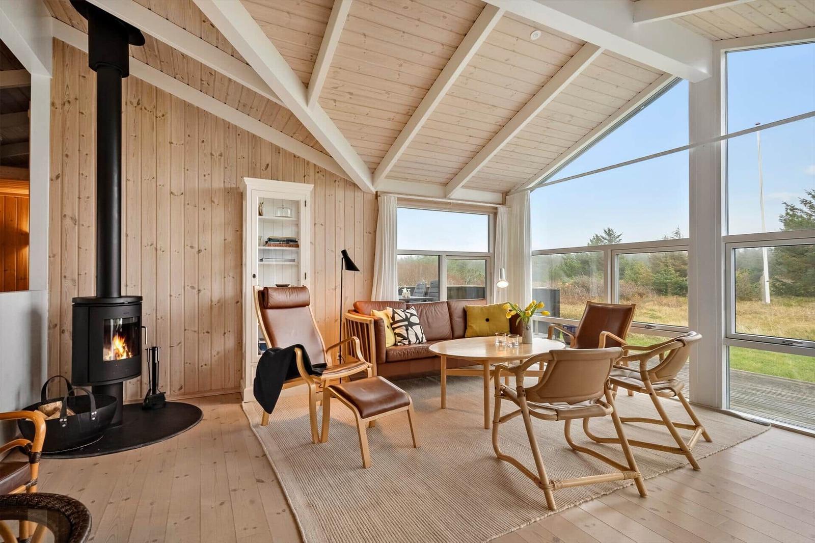Wohnraum mit Kamin, Sofa und Stühlen. Große Fenster bieten Blick auf die Landschaft.
