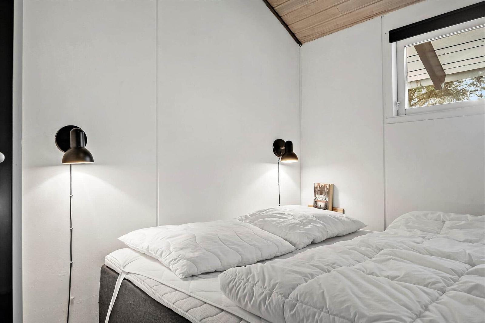 Schlafzimmer mit Bett, weißen Kissen und Decke, zwei Wandlampen und kleinem Fenster.
