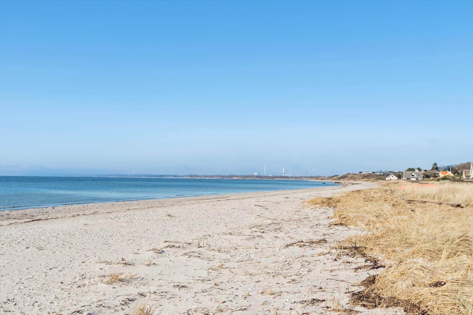 Strand mit Sand, trockener Vegetation und Blick aufs Meer und Ferienhäuser.