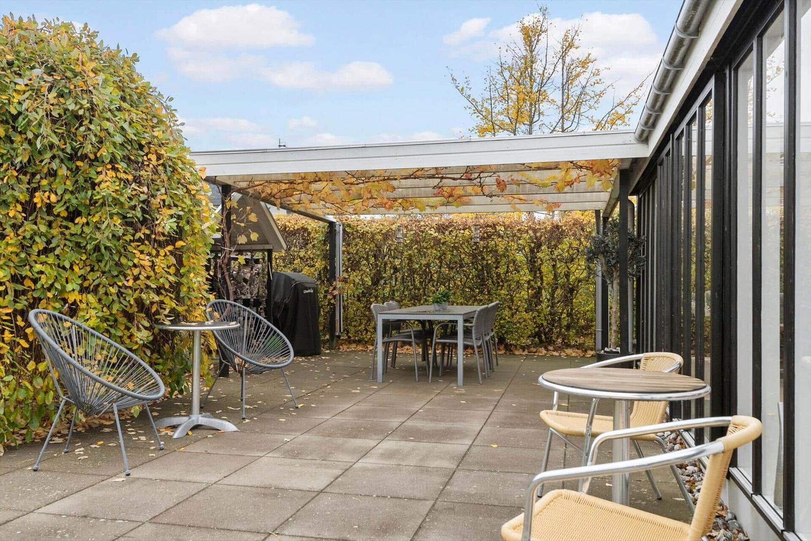 Terrasse mit Tischen, Stühlen und Pergola unter Herbstlaub.