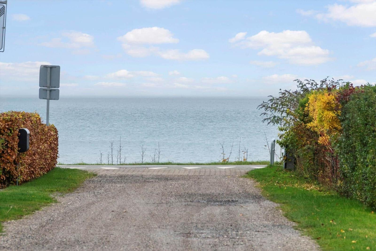 Grauer Kiesweg führt zum Ufer. Grünflächen und Sträucher säumen den Weg. Im Hintergrund erstreckt sich das Meer.