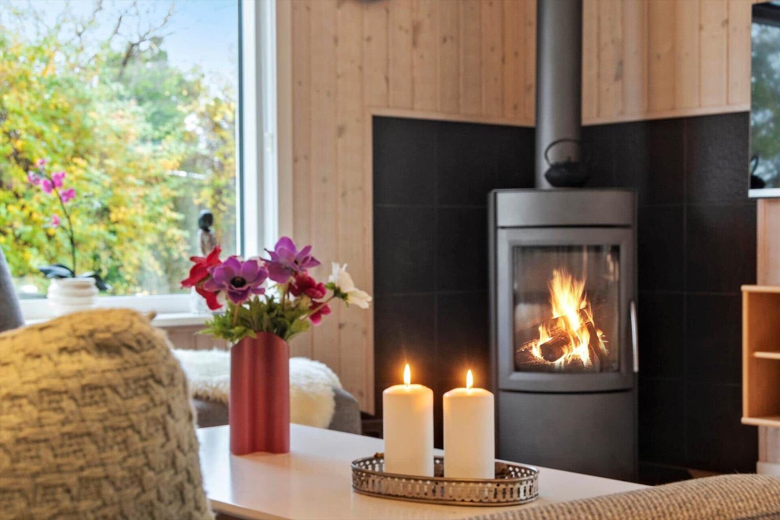 Ein Kamin mit brennendem Holz, zwei Kerzen und Blumen vor einem Fenster.