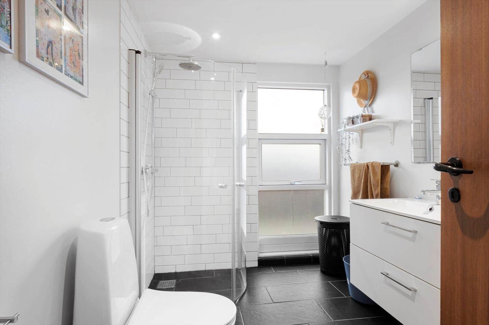 Badezimmer mit Dusche, Waschbecken und Toilette. Weiße Fliesen und dunkle Bodenfliesen.