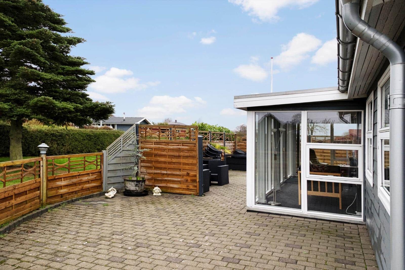Hinterhof mit Steinplatten, Holzgeländer und Glasveranda. Gartenmöbel und Pflanzen sind sichtbar.