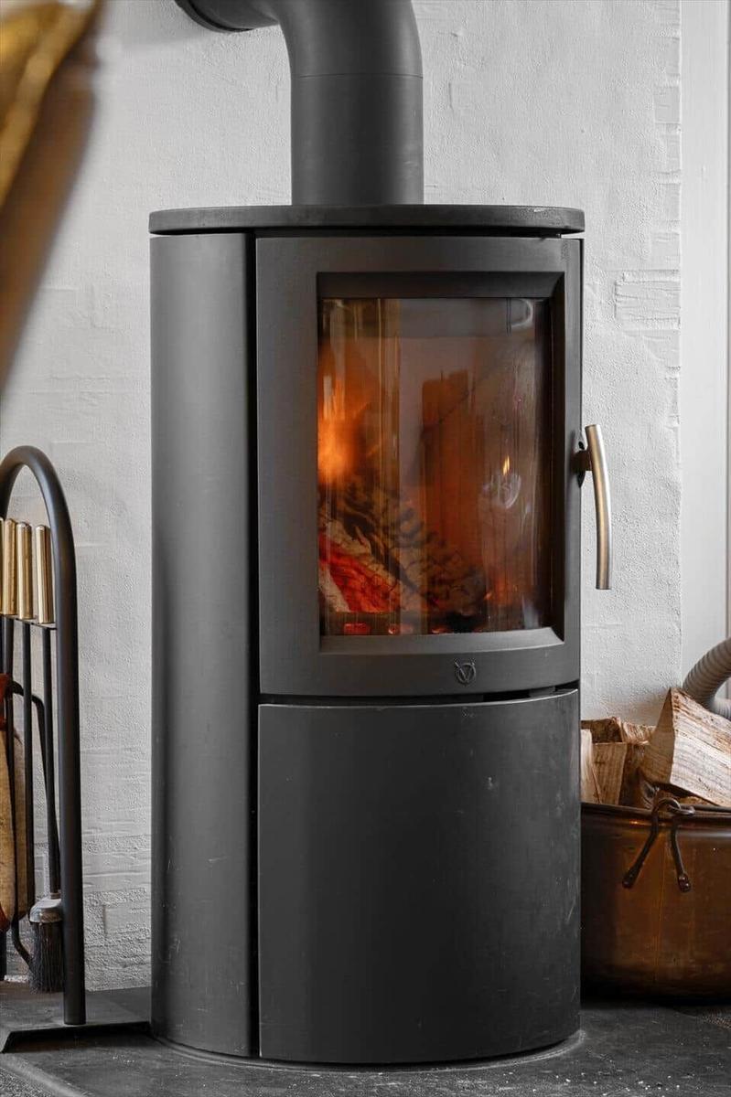 Schwarzer Kaminofen mit brennendem Holz, daneben Holzstapel und Feuerzeugständer.