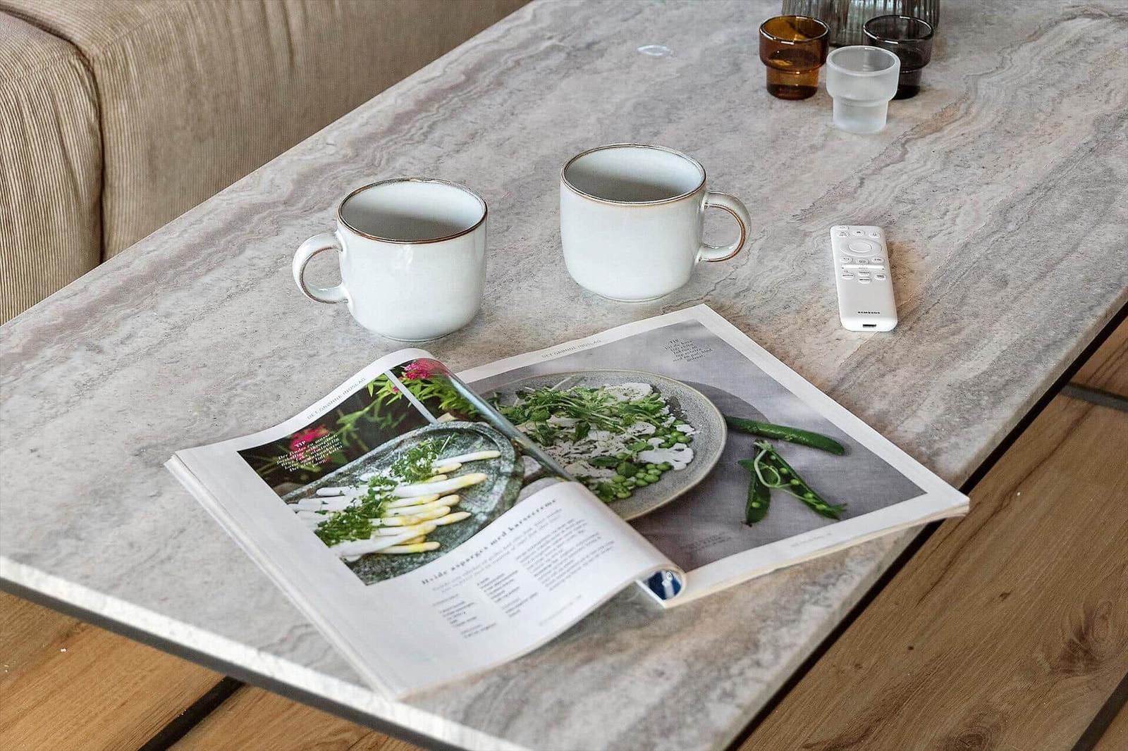 Tisch mit Tassen, Fernbedienung und geöffnetem Kochbuch.