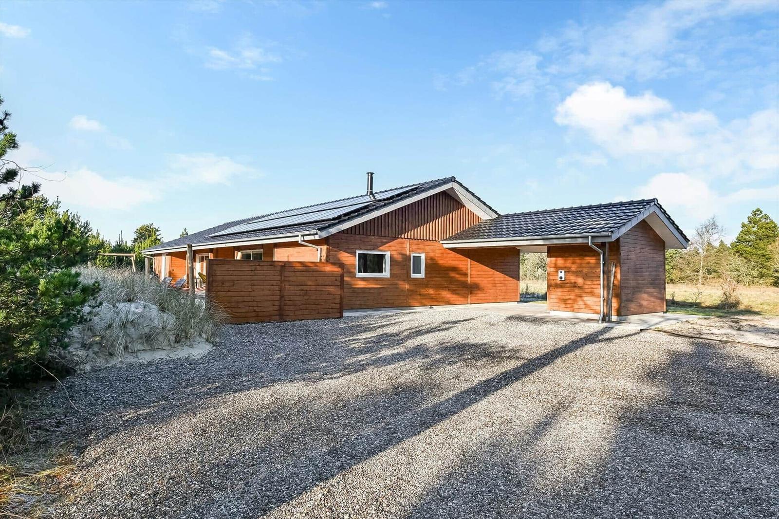 Wooden house with roof and garage on gravel ground. Surrounded by trees and sky.