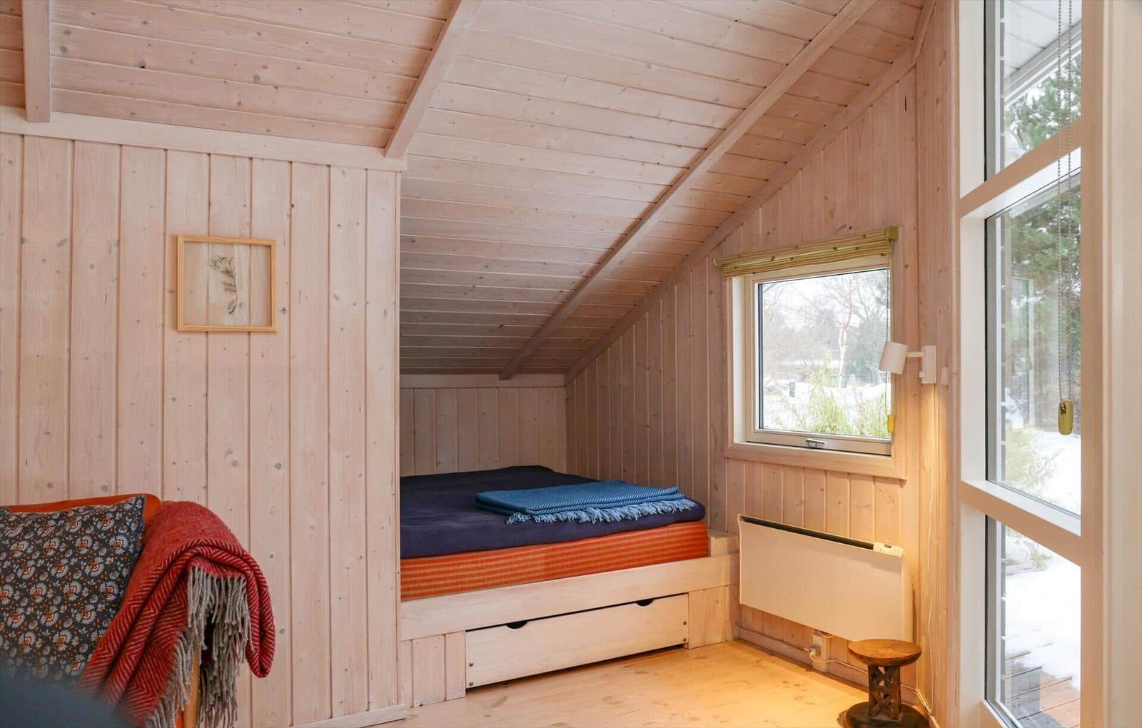 Schlafzimmer mit Holzwand, Bett und Fenster. Ein kleiner Holztisch steht daneben.